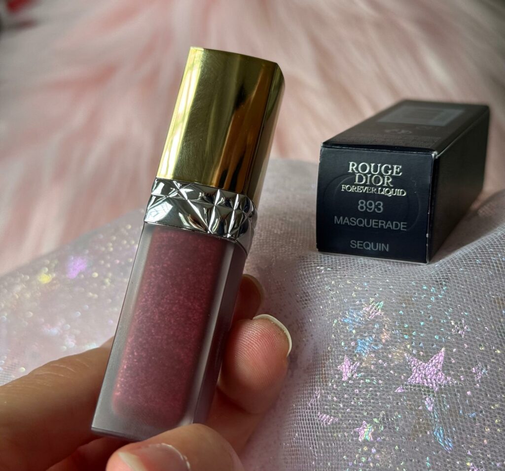 Nell'immagine si vede il rossetto Rouge Dior forever liquid nella tonalità 893 masquerade sequin. Il rossetto viene tenuto in mano e mostra tutti i suoi riflessi, sullo sfondo si vede la scatola.